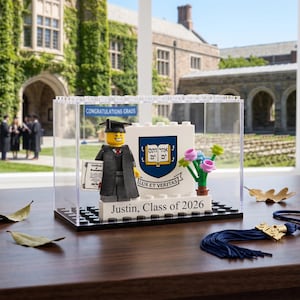 May include: A Lego graduation scene inside a clear display case. The scene includes a Lego figure in a graduation gown, a diploma, and a university emblem. The base reads "Justin, Class of 2026".