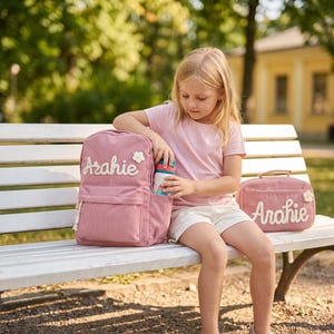 May include: A dusty pink corduroy backpack and a matching rectangular lunch bag, both featuring the name Arahie in white script lettering and a small white flower appliqu&eacute;. The backpack has a front pocket and side sleeve for a water bottle.