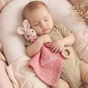 May include: A pink muslin security blanket featuring a crocheted bunny head with a small bow tie, a wooden ring attachment, and the name Sandra embroidered in white cursive script on the fabric. The blanket is soft, textured, and lightweight.