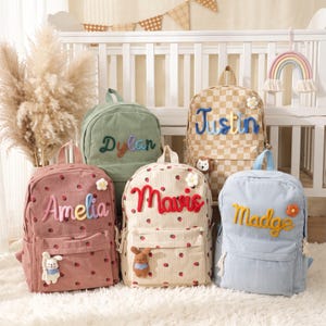 May include: Five corduroy backpacks in various colors including dusty pink, sage green, beige checkerboard, cream with strawberry prints, and light blue. Each bag features a personalized name in raised lettering and small decorative plush accents.