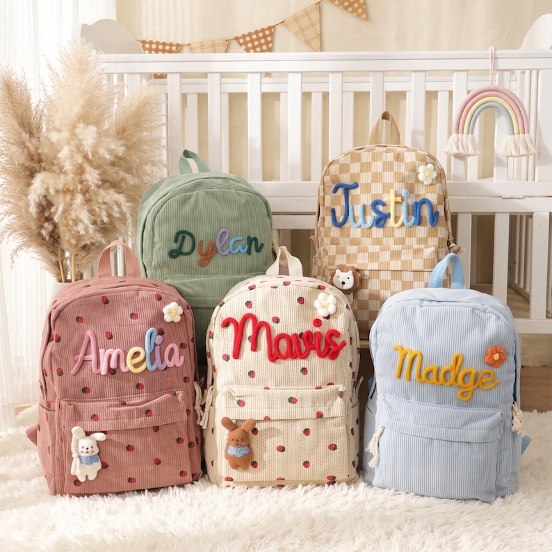 May include: Five corduroy backpacks in various colors including dusty pink, sage green, beige checkerboard, cream with strawberry prints, and light blue. Each bag features a personalized name in raised lettering and small decorative plush accents.