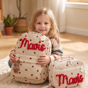 May include: A beige corduroy backpack and matching lunch bag featuring a red strawberry pattern, the name Mavis in red cursive embroidery, a small white flower appliqu&eacute;, and a brown plush rabbit charm attached to the backpack front pocket.