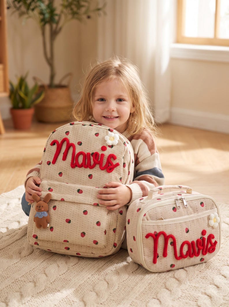 May include: A beige corduroy backpack and matching lunch bag featuring a red strawberry pattern, the name Mavis in red cursive embroidery, a small white flower appliqu&eacute;, and a brown plush rabbit charm attached to the backpack front pocket.