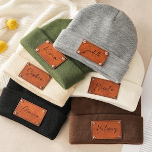 May include: A collection of personalized beanies in various colors, including black, brown, green, and gray. Each beanie features a brown leather patch with a name engraved on it. The names include Maria, Hilary, Kevin, Judith, Nicola, and Sophia.