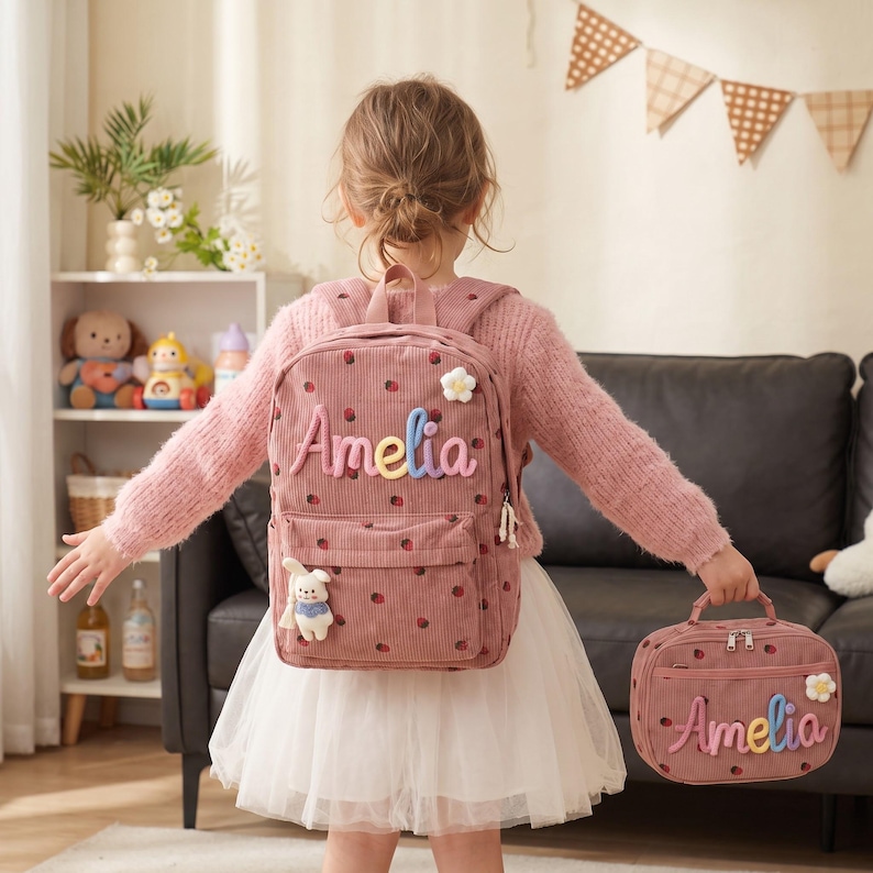 May include: A dusty pink corduroy backpack and matching lunch bag, both featuring a strawberry pattern, a white flower accent, and the name Amelia in colorful, raised, puffy lettering. A small plush rabbit charm hangs from the backpack zipper.