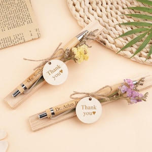 May include: Two bamboo pens with silver accents, each resting on a wooden base and tied with twine. Each pen is adorned with dried flowers and a wooden tag that says "Thank you". One tag reads "Ms. Sophia", the other "Dr. Arlen P".