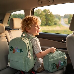 May include: A sage green corduroy backpack and a matching lunch bag, both featuring the name Dylan embroidered in colorful, raised letters. The backpack has white rope zipper pulls, and the lunch bag includes a small white flower charm.