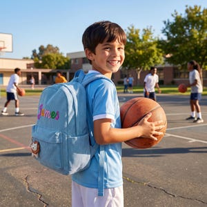 May include: A light blue corduroy backpack featuring the name James embroidered in colorful script on the front pocket, accompanied by a small lion-shaped zipper pull charm, worn by a child holding a brown basketball on an outdoor court.