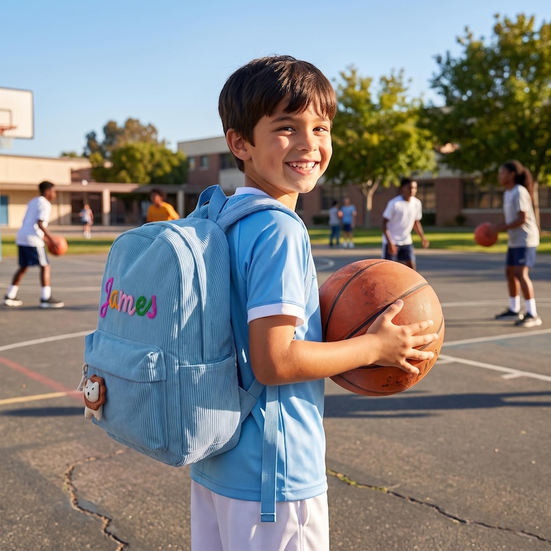 May include: A light blue corduroy backpack featuring the name James embroidered in colorful script on the front pocket, accompanied by a small lion-shaped zipper pull charm, worn by a child holding a brown basketball on an outdoor court.