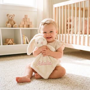 May include: A cream-colored plush security blanket featuring a stuffed rabbit head with long ears and a pink embroidered nose. The soft, square-shaped fabric is personalized with the name Adalia stitched in pink cursive script across the front.