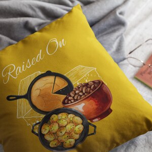 May include: Mustard yellow throw pillow with a watercolor illustration of Southern comfort food. The design includes a skillet of cornbread, a bowl of beans, and a pan of potatoes. The words "Raised On" are written in a script font.