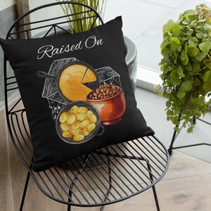 May include: Black throw pillow with a watercolor illustration of a skillet of cornbread, a pot of beans, and a skillet of potatoes. The words "Raised On" are written in white script across the top of the pillow.