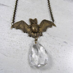 May include: A bronze-toned bat pendant necklace with a clear, teardrop-shaped crystal. The bat has outstretched wings and is suspended from a delicate chain. The crystal is faceted, catching the light. A gothic-inspired accessory.