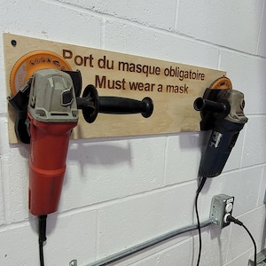 May include: Two angle grinders, one red and one blue, are mounted on a wooden sign that reads "Must wear a mask" in English and French. The grinders are attached to a white brick wall.