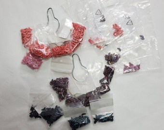 May include: Assortment of small plastic bags filled with colorful crescent-shaped beads. The bags are arranged on a white surface. The beads are in shades of pink, red, purple, and blue.
