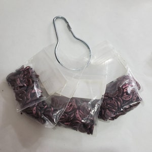 Pode incluir: Quatro sacos de pequenas contas de cor vermelha escura em forma de crescente. Os sacos est&atilde;o pendurados em um gancho de metal.
