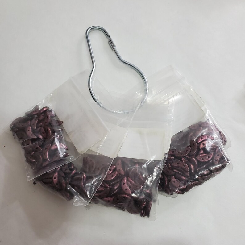 Pode incluir: Quatro sacos de pequenas contas de cor vermelha escura em forma de crescente. Os sacos est&atilde;o pendurados em um gancho de metal.