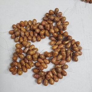 May include: A collection of teardrop-shaped beads in a muted mauve and bronze color palette. Each bead features a smooth, glossy finish with subtle, darker brown speckles distributed across the surface. The beads are strung on thin, clear filaments.