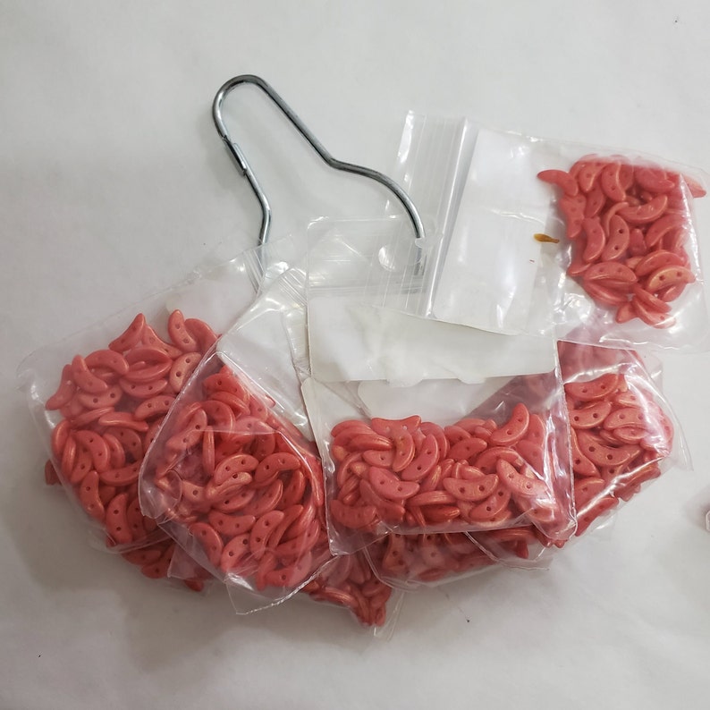 Pode incluir: V&aacute;rias sacolas de pequenas contas vermelhas em forma de meia lua. As contas t&ecirc;m um acabamento brilhante e met&aacute;lico.