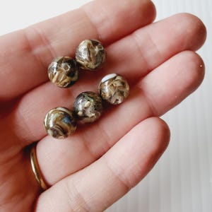 May include: A collection of six round beads with a marbled appearance. The beads have a mix of brown, tan, and white colors, with a glossy finish. The beads are small, each approximately 0.5 inches in diameter, and are likely used for jewelry making or crafts.