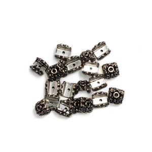 May include: A pile of silver-toned metal bead caps with a decorative design. The caps have a square shape with a small hole in the center.