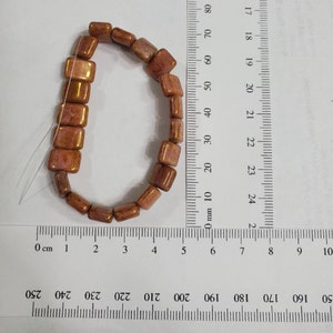 May include: A strand of 14 brown and pink square-shaped glass beads with a metallic sheen. The beads are strung on a clear thread and measure approximately 2.5 cm in length.