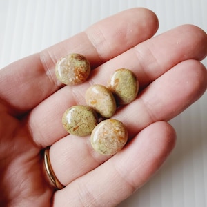 Peut inclure: Six pierres polies et rondes dans des tons de beige, marron et vert sont tenues dans une main ouverte. Les pierres ont une finition brillante et des motifs variés. Une bague en or est visible sur l'annulaire.