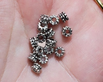 May include: A close-up of a pile of silver-toned metal beads with a decorative design. The beads are small and round, with a hole in the center for threading.