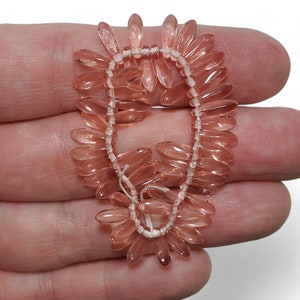 May include: A strand of pale pink glass beads, each bead is shaped like a long, thin oval with a pointed end. The beads are strung on a white thread.