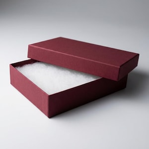 Puede incluir: Una caja de regalo rectangular de color burdeos con la tapa ligeramente abierta. El interior está forrado con un material blanco y esponjoso, probablemente para amortiguar objetos delicados. La caja está sobre un fondo blanco liso.