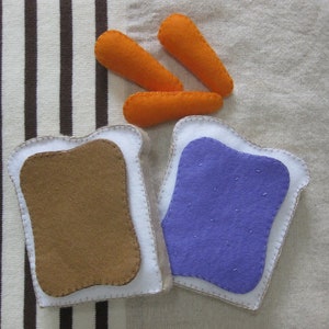 May include: Two felt pieces of bread with purple and brown felt filling. Three orange felt carrot sticks are on top of the bread.