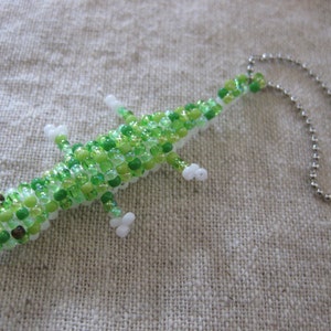 May include: A green and white beaded lizard charm with a silver chain attached. The lizard is made of small, round beads and has a white belly and green back.