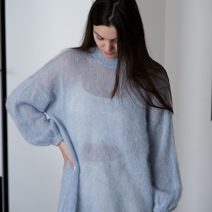 May include: A light blue, sheer knit sweater with long sleeves and a crew neck. The sweater is paired with white pants. The model is wearing the sweater and is standing in front of a white door.