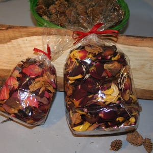 Könnte beinhalten: Zwei transparente Zellophanbeutel gefüllt mit getrockneten Rosenblättern, gebunden mit roten Bändern. Die Blütenblätter sind eine Mischung aus Rot, Gelb und Braun. Ein Holzbrett und ein grüner Korb mit Tannenzapfen befinden sich im Hintergrund.