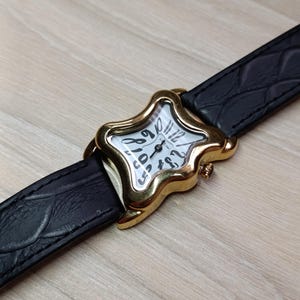 May include: A gold-toned wristwatch with a star-shaped face and a black leather band. The watch face is white with black numerals and hands. The band has a textured, crocodile-like pattern.
