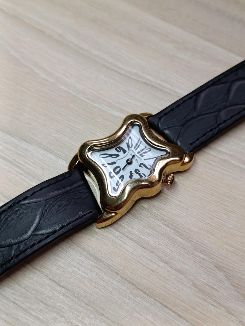May include: A gold-toned wristwatch with a star-shaped face and a black leather band. The watch face is white with black numerals and hands. The band has a textured, crocodile-like pattern.
