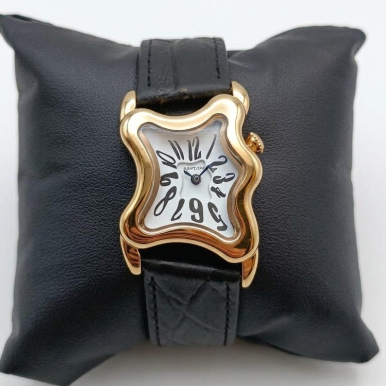 May include: A gold-toned wristwatch with a unique, melting design. The white watch face features black numerals and hands, set within a distorted square frame. The watch has a black leather band and is displayed on a black pillow.