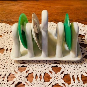 May include: A white ceramic plate rack holding several plates. The rack has a rectangular base with vertical dividers. Green and patterned plates are displayed. The rack sits on a white lace doily over a wooden surface.
