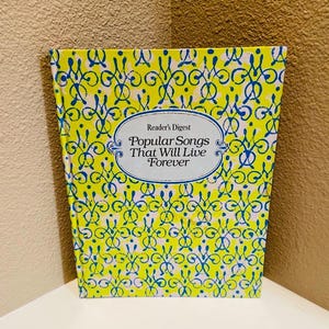 Może przedstawiać: Książka w stylu vintage zatytułowana "Popular Songs That Will Live Forever" wydawnictwa Reader's Digest. Książka ma jaskrawożółtą okładkę z niebiesko-białym wzorem. Tytuł znajduje się w białym owalu z niebieską obwódką.