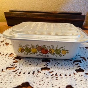 May include: A rectangular white ceramic baking dish with a clear glass lid. The side of the dish features a colorful botanical illustration of various vegetables and herbs, with the text L'Echalote printed in black script below the design.