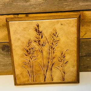 May include: A square wall art piece featuring a light tan ceramic tile with a dark brown embossed design of four stalks of wheat or grass, encased in a simple, medium-toned wooden frame with a smooth, clean finish.