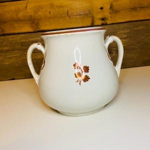May include: A white ceramic sugar bowl with two handles and a floral design. The rim and floral accents are a reddish-brown color. The sugar bowl is a vintage style.