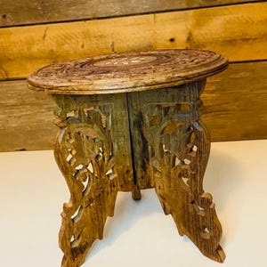 May include: A small, intricately carved wooden table with a round top and three ornate legs. The table is made of light brown wood and features detailed floral patterns. The table is approximately 12 inches in diameter and 10 inches tall.
