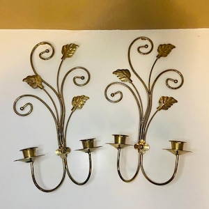 May include: Pair of vintage brass wall sconces. Each sconce features a floral and leaf design with two candle holders. The sconces are made of brass and have a warm, golden color. The overall design is elegant and decorative.
