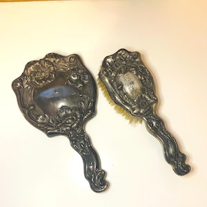 May include: A silver-toned hand mirror and a matching hairbrush set. Both items feature an ornate, raised floral relief pattern on their metallic handles and backs. The hairbrush has light-colored bristles visible along the edges of the frame.
