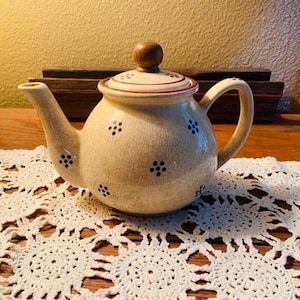 May include: A cream-colored ceramic teapot with a crackle glaze finish, featuring small dark blue floral patterns across the body and lid. The teapot has a rounded shape, a curved handle, a spout, and a small, round, light brown wooden knob on the lid.