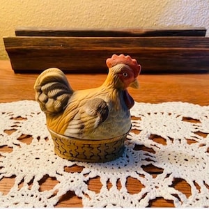 May include: A ceramic hen figurine with a red comb and wattle, tan and brown feathers, and a basket-weave base. The figurine is sitting on a white lace doily. The background includes a wooden box and a wooden table.