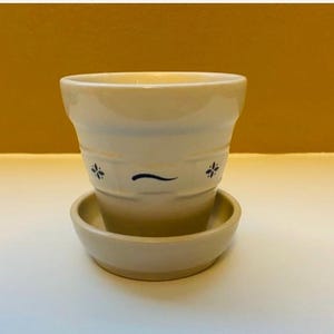 May include: A cream-colored ceramic flower pot with a matching saucer. The pot has a slightly flared shape with horizontal ridges and is decorated with small blue floral accents and a curved line. The saucer is round and shallow.