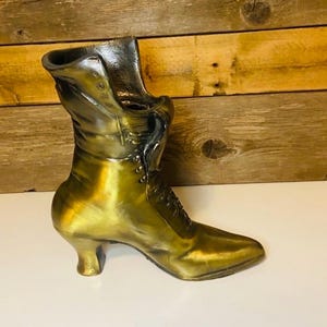 May include: A vintage-style, gold-colored boot-shaped vase. The boot features a pointed toe, a small heel, and button and lace details. The vase is made of metal and has a glossy finish. The background is a wooden wall.