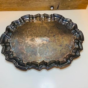 May include: Oval silver-plated tray with a decorative, scalloped edge. The tray has a tarnished appearance, showing signs of age and use. The tray is empty and sits on a white surface, with a wooden element in the background.